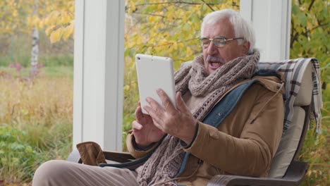 Ein-älterer-Mann-Sitzt-Auf-Einer-Bank-Im-Heimischen-Garten,-Während-Er-Mit-Einem-Tablet-über-Einen-Videoanruf-Spricht
