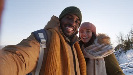 Happy-Young-Intercultural-Couple-In-Winterwear-Waving-At-The-Camera-Standing-Against-Blue-Sky-Over-Mountains-Covered-With-Snow-During-Chill-On-Winter