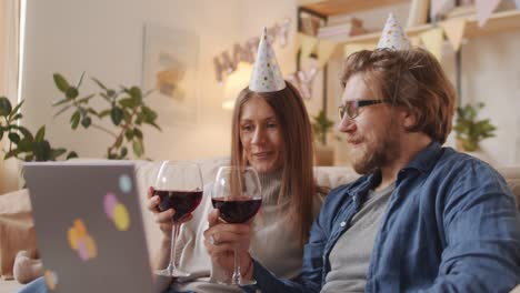 Couple-Celebrating-Birthday-Online-Wearing-Birthday-Hats-And-Toasting-With-Glasses-Of-Red-Wine