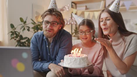 Parents-And-Daughter-Celebrating-Birthday-Wearing-Birthday-Hats,-The-Greeting-The-Camera-With-A-Birthday-Cake