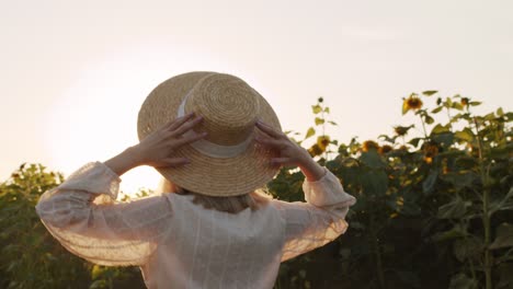Rear-View-Of-Beautiful-Young-Woman-In-Straw-Hat-And-White-Country-Style-Dress-Smiling-And-Dancing-In-Sunflowers-Field-At-Sunset