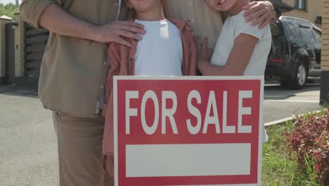 Family-With-Two-Children-Looking-At-Camera-In-The-Garden-At-Home-Holding-A-For-Sale-Signboard-To-Move-To-Another-Home