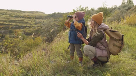 Young-Female-Backpacker-Squatting-By-Her-Adorable-Little-Son-With-Binoculars-During-Weekend-Trip-In-Natural-Environment