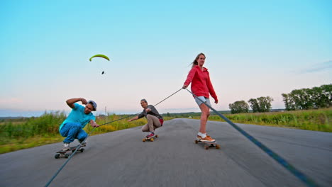 Front-View-Of-Two-Boys-And-A-Girl-Skateboarding-Holding-On-To-A-Rope-On-The-Road,-In-The-Sky-A-Parachutist-Flying