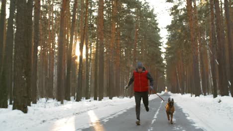 Man-Dressed-In-Winter-Sportswear-Running-With-His-Dog-On-The-Road-In-The-Snowy-Forest