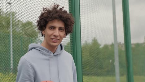 Young-Smiling-Active-Male-Basket-Player-In-Gray-Sweatshirt-Holding-Ball-While-Standing-On-Playground