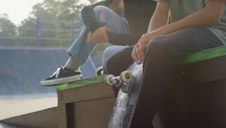 Young-Man-Sitting-With-A-Friend,-Spinning-A-Skateboard-Wheel-In-Skate-Park