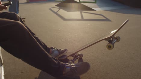 Camera-Focuses-On-Young-Man-Sitting-On-Skatepark,-While-His-Friend-Jump-On-Skateboard