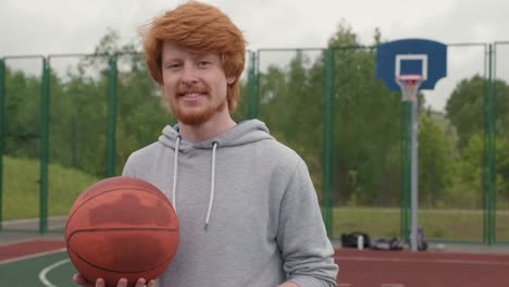 Young-Smiling-Redhead-Athlete-With-Sweatshirt-Holding-Ball-For-Playing-Basketball-In-Basketball-Court