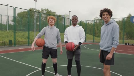 Three-Friends-With-Basketballs-On-Basketball-Court,-Smiling-And-Looking-At-Camera