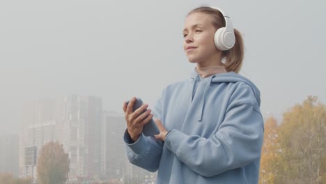 Young-Serious-Sportswoman-In-Blue-Hoodie-With-Headphones-Looking-At-Smartphone-Before-Training-In-Forest-Path