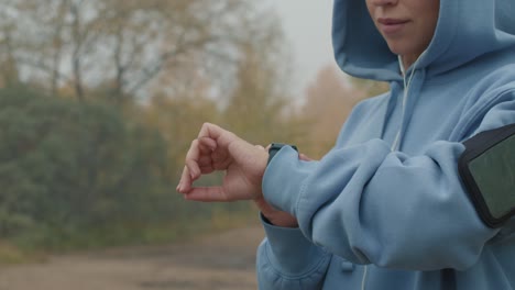 Young-Serious-Sportswoman-In-Blue-Hoodie-Looking-At-Smartwatch-Before-Training-In-Forest-Path