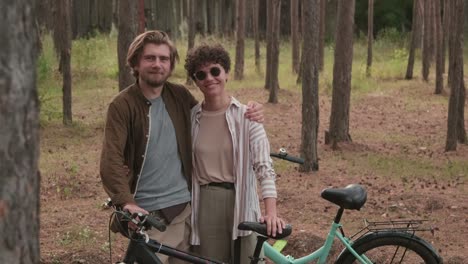 Young-Affectionate-Man-Embracing-His-Happy-Wife-In-Sunglasses-While-Both-Standing-With-Their-Bicycles-In-Rural-Environment
