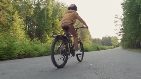 Vista-Trasera-De-Un-Niño-Montando-En-Bicicleta-En-Un-Camino-Forestal-Mientras-Otras-Personas-Corren