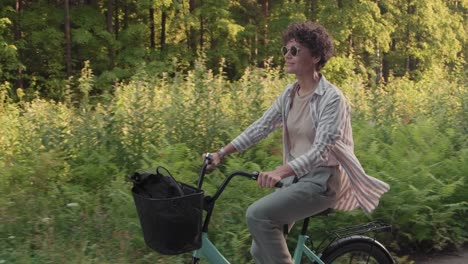 Junge-Glückliche-Frau-In-Sonnenbrille-Und-Lässiger-Kleidung,-Die-Mit-Dem-Fahrrad-Auf-Einem-Waldweg-Fährt