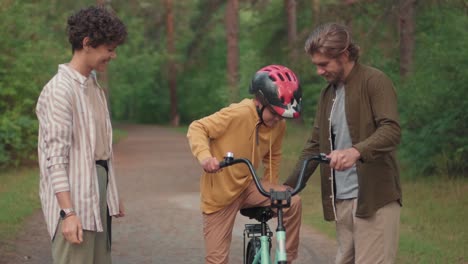 Mother-Puts-The-Helmet-On-Her-Son-While-His-Father-Holds-His-Bike,-Then-Child-Rides-His-Bike-In-The-Forest