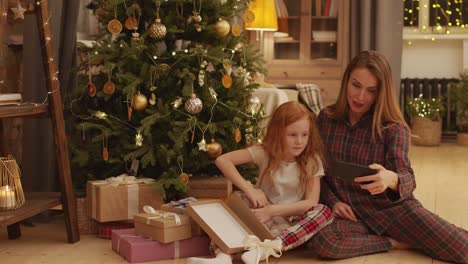 Glückliche-Junge-Mutter-Und-Entzückende-Kleine-Tochter-Im-Pyjama,-Die-Selfie-Durch-Geschmückten-Weihnachtsbaum-Machen,-Während-Sie-Auf-Dem-Boden-Des-Wohnzimmers-Sitzen