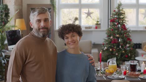 Couple-Looks-At-The-Camera-And-Smiles-In-Living-Room-With-Table-And-Christmas-Decorations