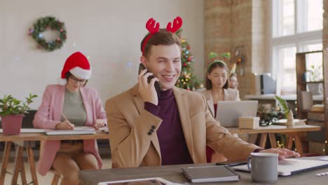 Junger-Seriöser-Geschäftsmann-In-Weihnachtskopfbedeckungen,-Der-Am-Telefon-Spricht,-Während-Er-Die-Arbeit-Plant,-Die-An-Heiligabend-Vor-Dem-Laptop-Sitzt