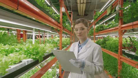Young-Greenhouse-Female-Worker-In-White-Coat-And-Eyeglasses-Holding-Clipboard-While-Looking-At-Camera-Inside-Vertical-Farm