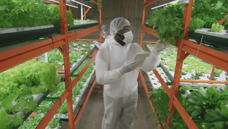 Young-Male-Agroengineer-In-Protective-Eyeglasses-And-Coveralls-Holding-A-Tablet-And-Walking-On-The-Hallway-Looking-Seedlings-Of-Green-Lettuce-In-Modern-Vertical-Farm-1