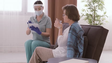 Senior-Woman-Drinking-Water-While-Talking-To-Female-Doctor-In-Medical-Mask-And-Protective-Screen-Using-A-Table