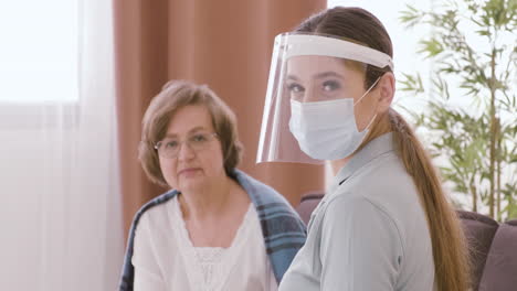 Doctora-Con-Máscara-Facial-Y-Pantalla-Protectora-Sentada-Con-Una-Anciana-Paciente-Mirando-La-Cámara