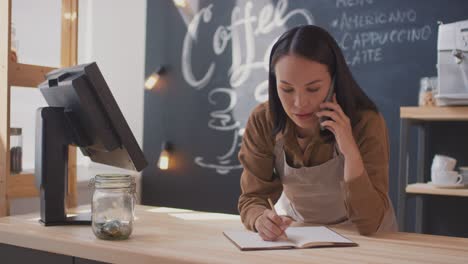 Female-Waitress-Taking-Notes-And-Talking-On-The-Phone-On-The-Counter-In-A-Coffee-Shop