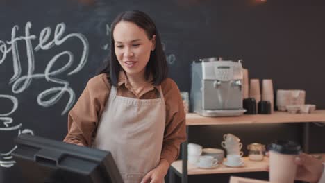 Waitress-In-Apron-Sells-A-Customer-A-Takeaway-Coffee-And-Food-Behind-The-Counter-In-A-Coffee-Shop