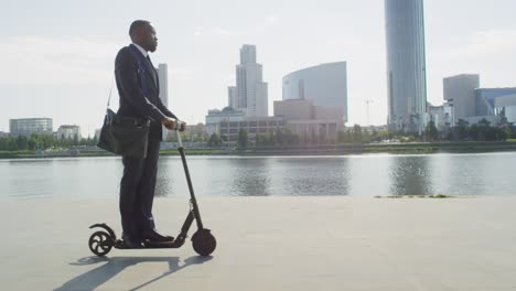 Young-Serious-Businessman-On-Electric-Scooter-By-Riverside-Against-Tall-Modern-Buildings