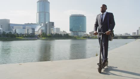 Young-Serious-Businessman-On-Electric-Scooter-By-Riverside-Against-Tall-Modern-Buildings-1