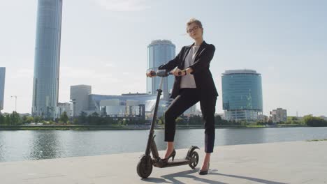 Young-Elegant-Businesswoman-Standing-On-Electric-Scooter-By-Riverside-Against-Modern-Buildings-On-Sunny-Day