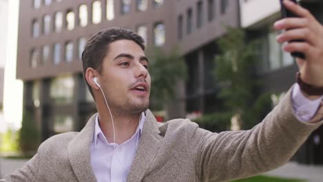 Young-Businessman-With-Earphones-Looking-At-Smartphone-Screen-During-Communication-In-Urban-Environment