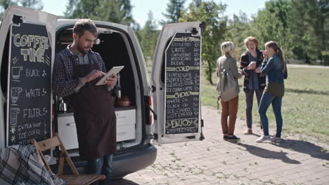 Coffee-Truck-Worker-Leaning-On-The-Van-While-Using-A-Tablet,-Group-Of-People-Talk-And-Drink-Coffee-In-The-Background