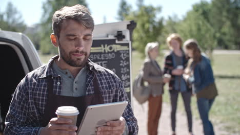 Coffee-Truck-Worker-While-Using-A-Tablet-And-Drinking-Coffee,-Group-Of-People-Talk-And-Drink-Coffee-In-The-Background