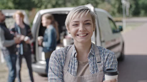 Coffee-Truck-Female-Worker-Looking-At-Camera-Smiling-And-Holding-A-Coffee,-Group-Of-People-Talk-And-Drink-Coffee-In-The-Background