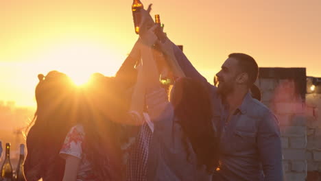 Group-Of-Friends-Enjoying-A-Party-On-A-Terrace-At-Sunset,-Hold-Drinks-With-Arms-Raised