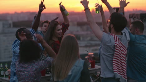 Group-Of-Friends-Enjoying-A-Party-On-A-Terrace-At-Sunset,-Hold-Drinks-With-Arms-Raised-1