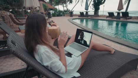 Woman-Wearing-White-Shirt-And-Drinking-From-A-Coconut-On-Video-Call-With-An-Coworker-While-Sitting-In-The-Hammock-In-Front-Of-The-Pool-In-Resort