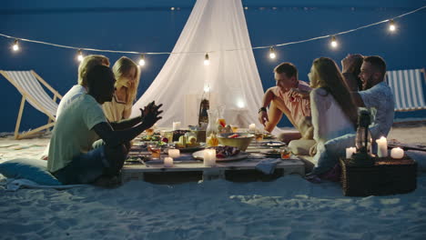 Group-Of-Friends-Having-Dinner-Sitting-On-The-Beach,-Smiling-And-Toasting,-With-A-Low-Table-And-A-Tent