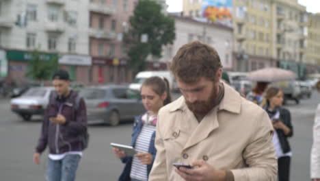 Young-Blond-Man-In-Raincoat-Walking-Down-The-Street-And-Using-Smartphone,-In-The-Background-People-Walking