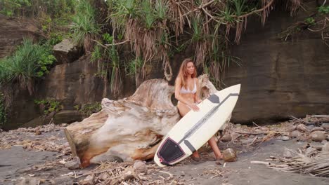 Concentrated-Beautiful-Girl-Leaning-Against-A-Trunk-On-A-Wild-Beach-With-Her-Surfboard-Ready-To-Surf
