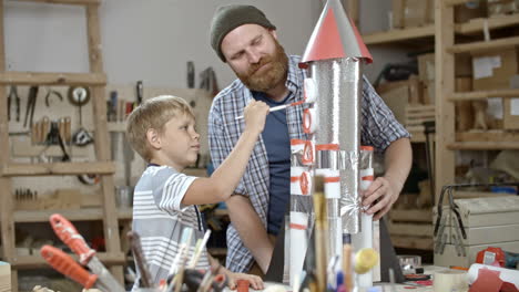 Kid-Malt-Mit-Hilfe-Seines-Vaters-In-Der-Bastelwerkstatt-Eine-Handgefertigte-Weltraumrakete