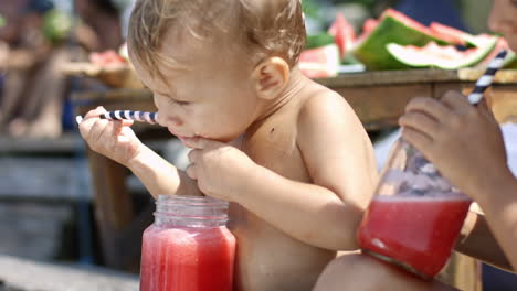 Camera-Focuses-On-A-Wet-Boy-Without-Clothes-On-The-Beach-While-He-Drinks-A-Watermelon-Juice-With-A-Straw-Accompanied-By-His-Sister