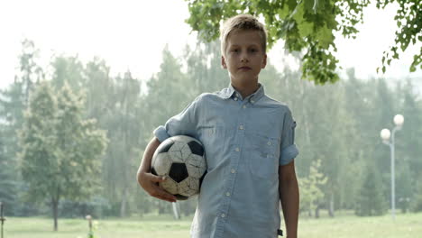 Blond-Boy-In-Blue-Shirt,-Kicks-Soccer-Ball,-Catches-It-With-His-Hands-And-Looks-At-The-Camera-In-The-Park