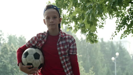 Boy-In-Blue-Cap-And-Plaid-Shirt-Bends-Down-To-Catch-Soccer-Ball-In-Park-And-Looks-At-Camera