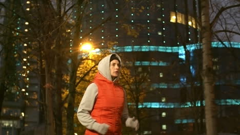 Camera-Focuses-On-A-Woman-In-Sportswear-And-Hood-Running-In-The-City-At-Night-In-Winter