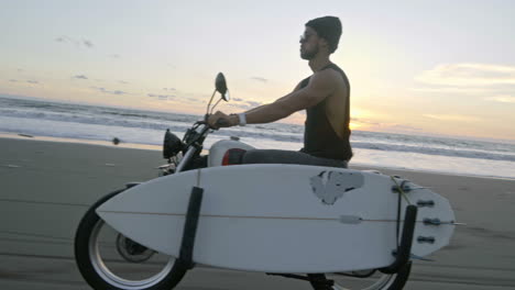 Hombre-Con-Sombrero-De-Lana-Y-Gafas-De-Sol-Montando-Una-Motocicleta-En-La-Playa-Con-Una-Tabla-De-Surf-A-Su-Lado