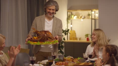 Gray-Haired-Man-Brings-The-Cooked-Turkey-To-The-Table-Where-The-Family-Is-Sitting-To-Celebrate-Thanksgiving