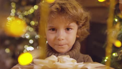 Happy-Child-With-Green-Eyes-And-Turtleneck-In-The-Foreground,-Looks-At-The-Camera-While-Holding-A-Gift-At-Christmas